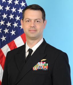 Commander Day has short dark hair, and brown eyes, is wearing a uniform jacket with many medals, and stands in front of an American flag and light blue background.