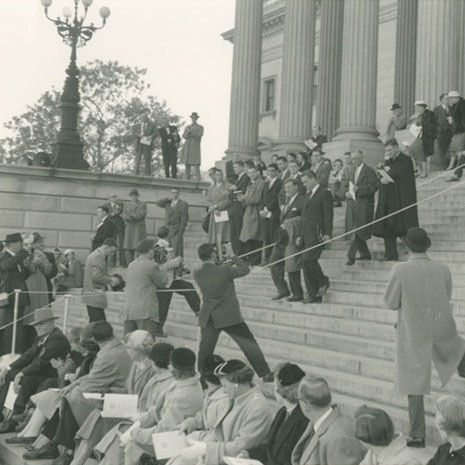 Black and white image of people gathered on steps of South Carolina State House. People in foreground seated on lower steps. In background, a group of people walk down steps, people with cameras photograph them from below.