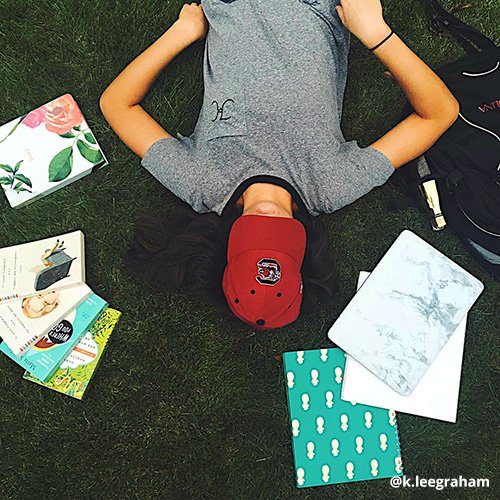 Student lays on ground, Carolina cap covering face, surrounded by books.