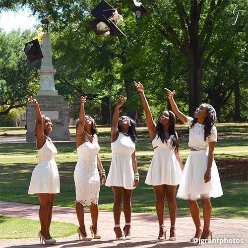 Five female students stand in white dresses throwing graduation caps in the air, Maxcy Monument behind them.