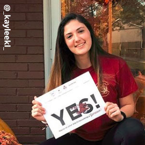 Image provided by @_kayleekl_ of a young woman with long brunette hair in a garnet USC t-shirt, smiling and sitting in front of a glass door holding an acceptance envelope that says, Yes! 