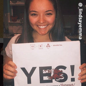 Image provided by @_lindsayemma of a young woman with straight, dark brown hair smiling and holding an acceptance envelope in front of her that says, Yes!
