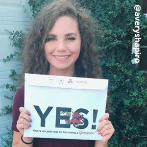 Image provided by @averyshpiro of a young woman with curly, light-brown hair standing outside in a garnet shirt and smiling, while holding an acceptance envelope that says, Yes!