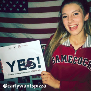 Image provided by @carlywantspizza of a young, blonde woman standing in front of an American flag and wearing a Gamecock pullover with a striped collar. She is smiling and holding an acceptance envelope that says, Yes!