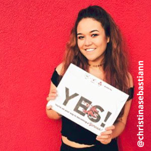 Image provided by @christinasebastiann of a young woman with long, brown hair in front of a red background. She is wearing an off-the-shoulder black top and smiling while she holds an acceptance envelope that says, Yes!
