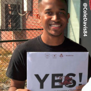 Image provided by @ColeDavis84 of a young man in front of a tall, chain-link fence. He is wearing a black t-shirt and smiling as he holds an acceptance envelope that says, Yes!