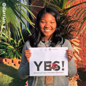 Image provided by @darrian_g of a young woman with black hair, smiling and holding an acceptance envelope that says, “Yes!” She is standing outside on a green lawn wearing a grey shirt.