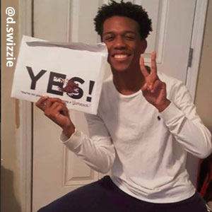 Image provided by @d.swizzle of a young man squatting in front of a white door, making a peace sign with his left hand. He is smiling happily and holding an acceptance envelope in his right hand that says, Yes!