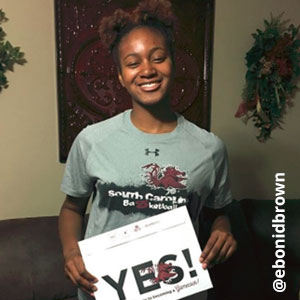 Image provided by @ebonidbrown of a young woman with her hair pulled up. She is smiling and wearing a grey South Carolina Basketball t-shirt, while she holds an acceptance envelope that says, Yes!
