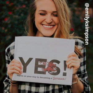 Image provided by @emilyksimpson of a young, blonde woman grinning with her eyes closed and holding an acceptance envelope under her chin that says, Yes!