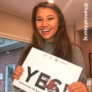 Image provided by @hannahlauraj of a young woman with long, brown hair wearing a grey shirt. She is smiling and holding an acceptance envelope that says, Yes!