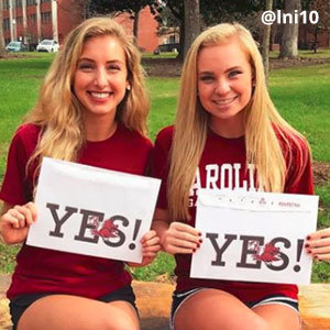 Two blonde girls are sitting in front of a green lawn in shorts and garnet t-shirts, smiling and holding acceptance envelopes that say, Yes! Image provided by @lni10.