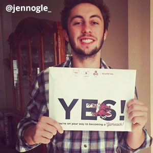 Image provided by @jennogle_ of a young, man smiling and holding an acceptance envelope that says, Yes! He is bearded with brown hear.