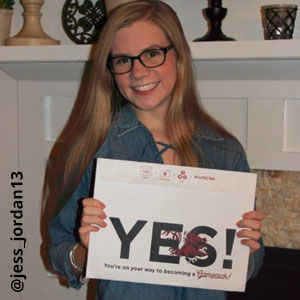 Image provided by @jess_jordan13 of a young, woman with long blonde hair standing in front of a decorated mantle and smiling. She wears black-framed glasses and holds an acceptance envelope that says, Yes!