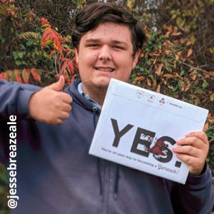 Image provided by @jessebreazeale of a young man with brown hair wearing a grey, zippered sweatshirt. He is standing outside, smiling and giving a thumbs up with his right hand while also holding an acceptance envelope that says, Yes! in his left hand.