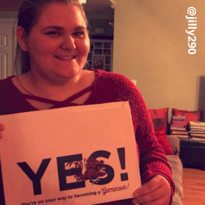 Image provided by @jilly290 of a young woman with her hair pulled back. She is wearing a red, long-sleeved sweater and smiling while she holds an acceptance envelope that says, Yes!
