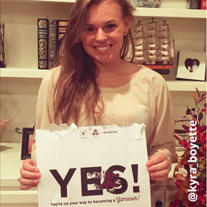 Image provided by @kyra_boyette of a young, smiling woman with long hair. She stands in front of a bookcase holding an acceptance envelope that says, Yes!
