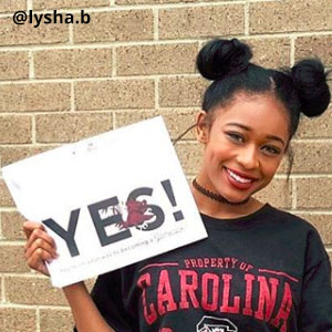 Image provided by @lysha.b of a young woman with her hair pulled up into puffs and wearing a black t-shirt that says Property of Carolina. She is standing in front of a brick wall and grinning while she holds an acceptance envelope that says, Yes! in her right hand.