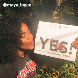 Image provided by @maya_logan of a young woman holding an acceptance envelope that says, “Yes!” and blowing it a kiss. She is outside, wearing a black t-shirt that says, Carolina Gamecocks.