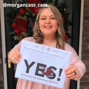 Image provided by @morgancass_cass of a young, woman with curled blonde hair and dimples. She stands in front of a wreath with red flowers, wearing a pink blouse. She smiling while she holds up an acceptance envelope that says, Yes!