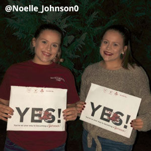 Image of two Caucasian girls who look very similar standing outside at night, smiling and holding acceptance envelopes that say, Yes! They both wear their hair pulled back and red lipstick. One wears a short-sleeved garnet t-shirt and the other wears a brown sweater that ties at the waist. Image provided by @Noelle_Johnson0
