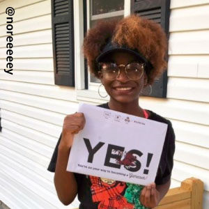 Image provided by @noreeeeey of a young girl wearing a sun visor, glasses and hoop earrings. She is standing in front of a white house with black shutters, smiling and holding an acceptance envelope that says, Yes!