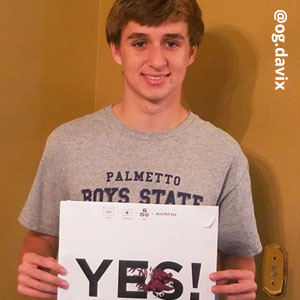 Image provided by @og.davix of a young, man wearing a grey t-shirt that says “Palmetto Boys State.” He is smiling and holding an acceptance envelope that says, Yes!