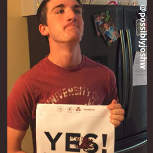 Image provided by @possiblyjoshw of a young, man in a red t-shirt standing in front of a black refrigerator. He is holding an acceptance envelope that says, Yes!
