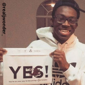 Image provided by @realjwonder_ of a young man in glasses and a hooded, white Adidas sweatshirt. He has a black hat turned to the back. He is  grinning proudly and holding an acceptance envelope that says, Yes!
