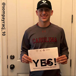 Image provided by sompayracj_10 of a young man wearing a grey shirt that says Carolina. He is smiling and holding an acceptance envelope that says, Yes!