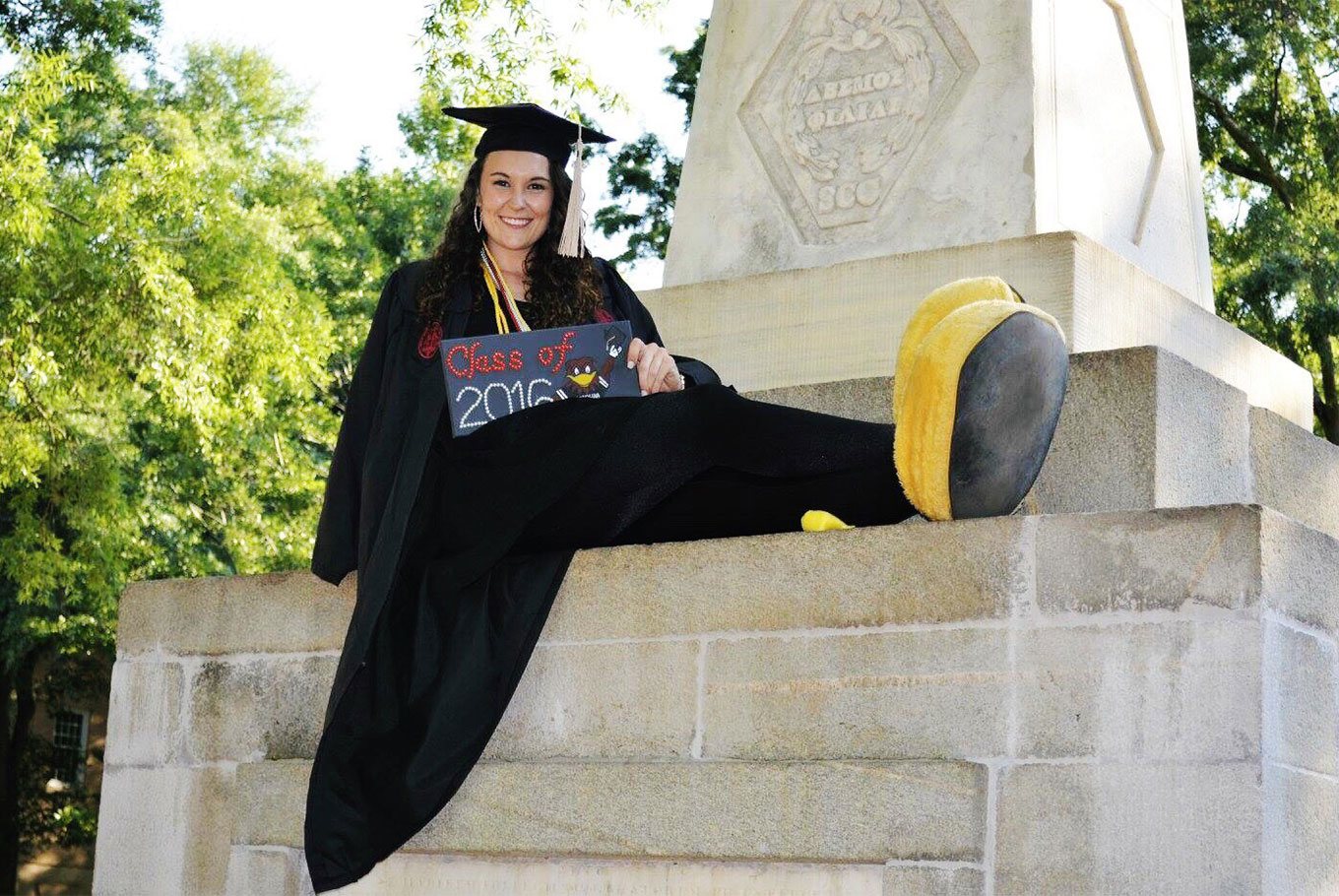 Graduating female senior poses on the Maxcy monument with her Cocky mascot feet on.