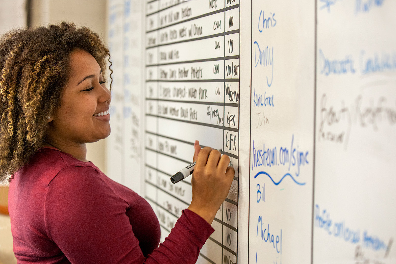 A journalism student smiles while filling in the day's television news production schedule on a dry erase board.
