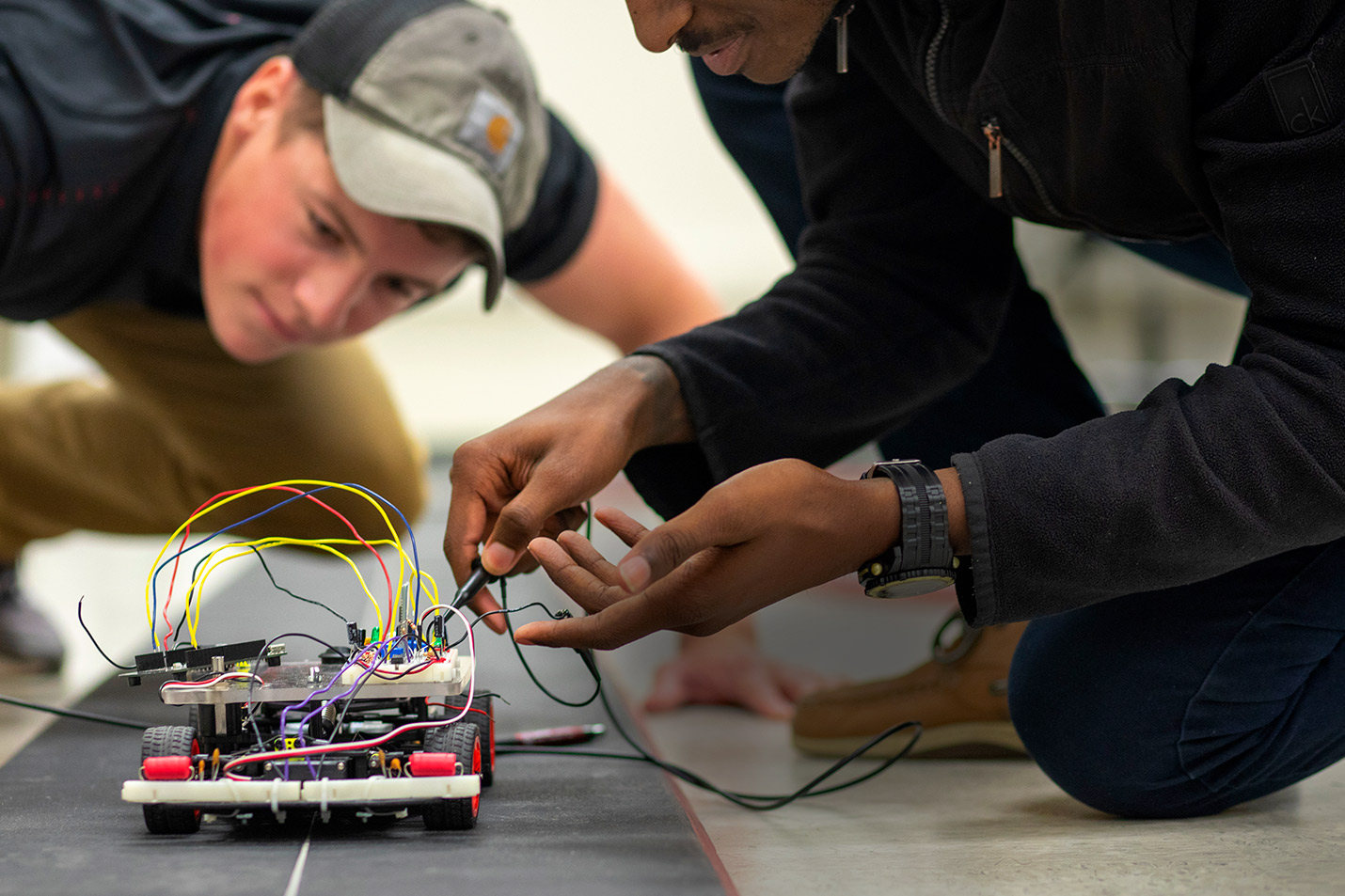Two students kneeling in the floor and working together on a small robotics project that has colorful wires and wheels.
