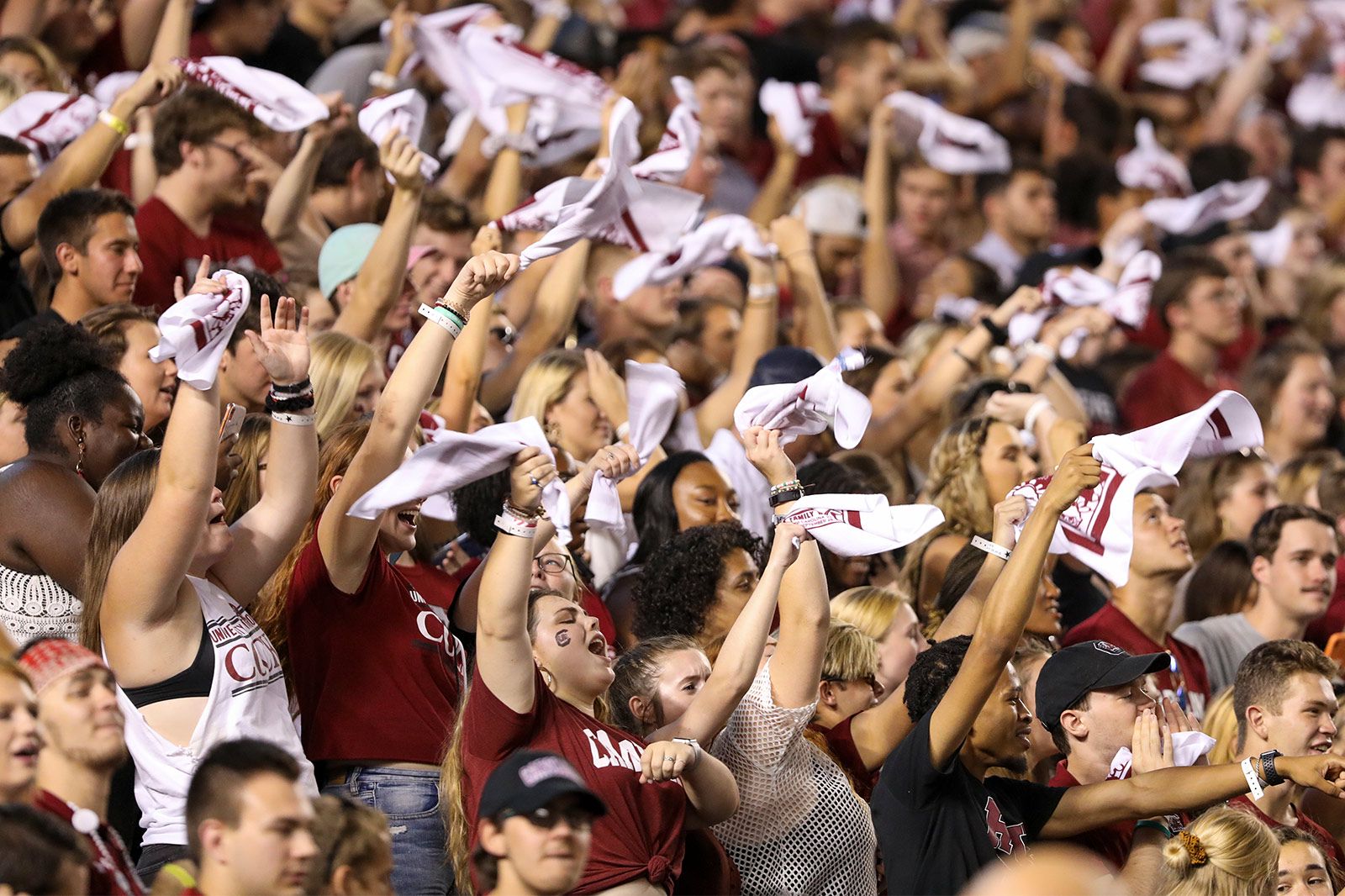 Large crowd of Gamecock students cheering in the stands at Williams Brice Stadium with arms raised, twirling towels in the air.