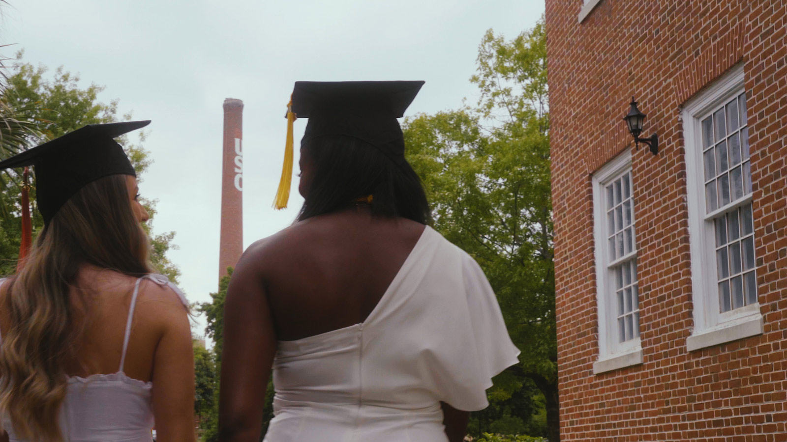 A view of two students from the back wearing graduation caps smiling at each other walking down a path on campus with the USC smokestack in the distance.