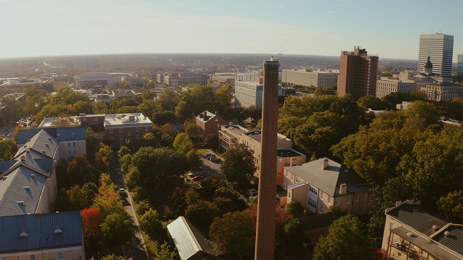 Aerial view of campus with rooftops, trees the smokestack in foreground and city of Columbia skyline off in the distance.