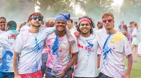 Four students at a Color Run wearing white shirts splattered with colored chalk posing for the camera with their arms around each other.