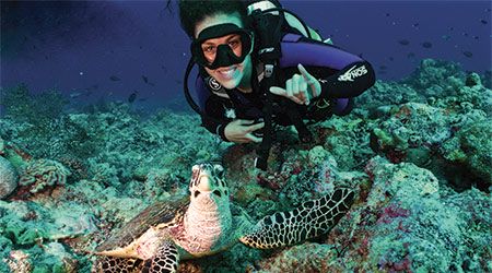 Student underwater scuba diving smiling for the camera with a sea turtle and giving a spurs up gesture with her hand.