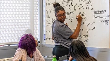 Student writing organic chemistry formulas on a whiteboard turned around looking at two other students sitting at a table.