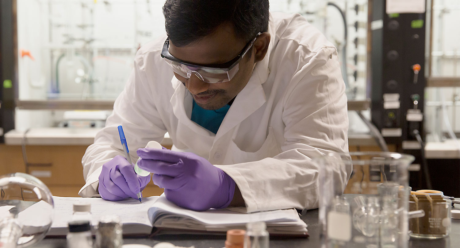 Graduate student leans over a notebook taking notes while holding a test tube in a lab setting. 