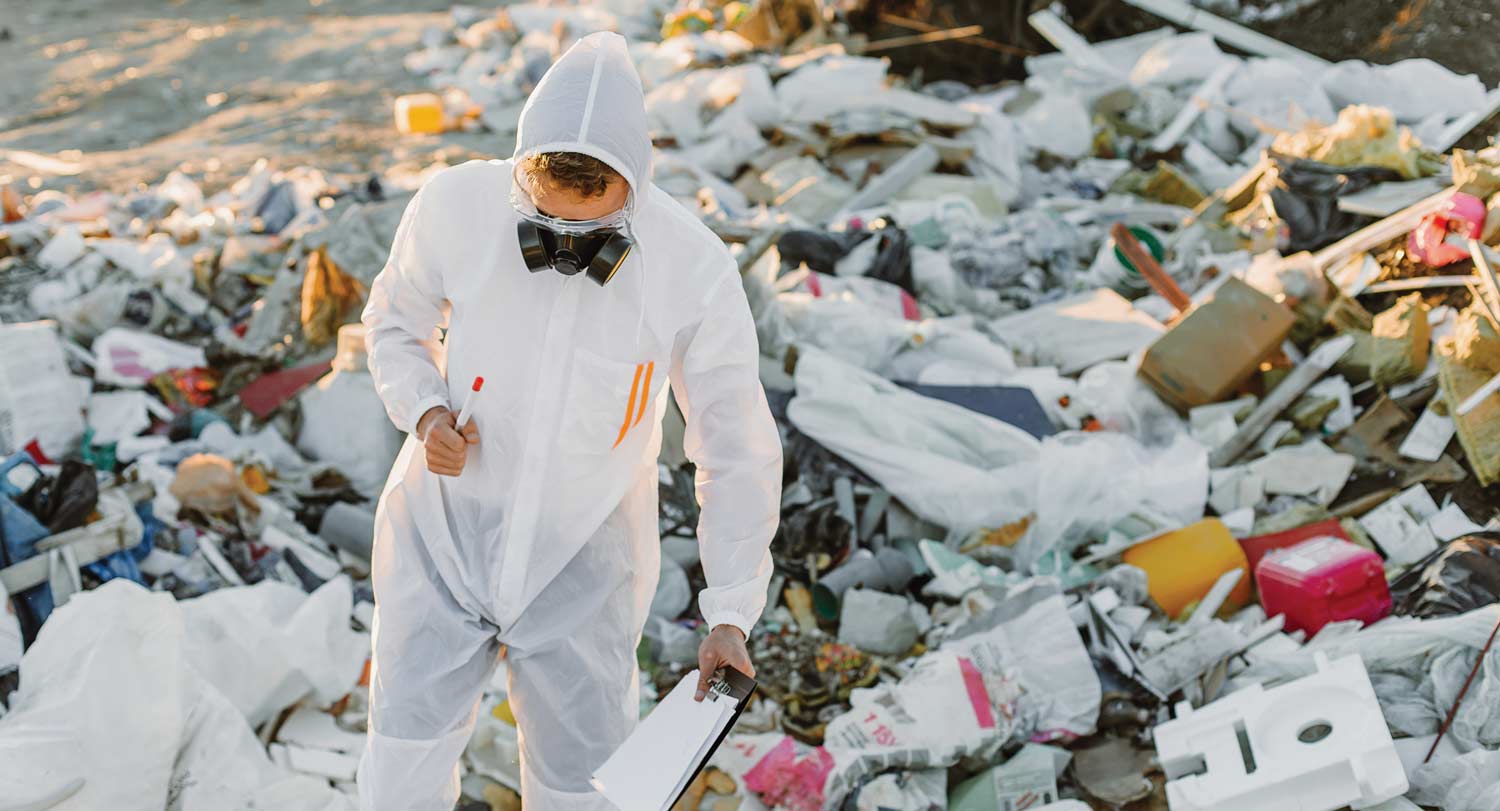 A photo of a scientist in a landfill wearing protective gear and carrying a clipboard.
