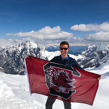 Student holding a Gamecock flag on the top of a snow covered mountain.