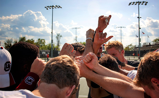 Students on a tennis court in a huddle with their arms raised together.