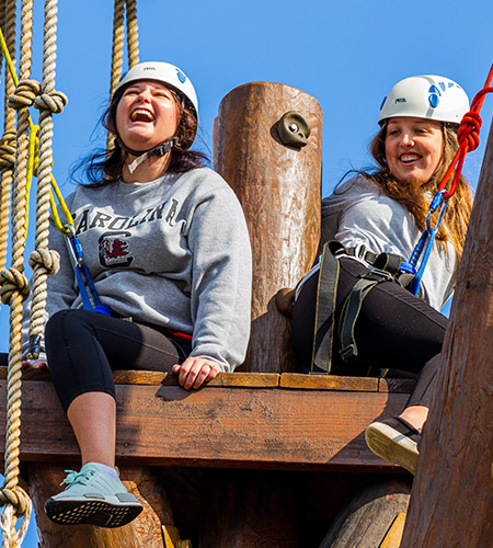 Students wearing harnesses and helmets sitting at the top of the ropes course laughing.