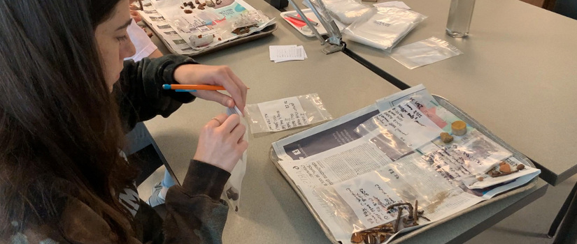 A student sits at a table tagging and bagging various artifacts.