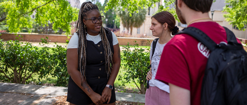 Three students stand outside in the area near the library. They are happy and chatting, with one facing away from the camera.