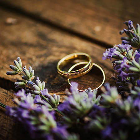 Two wedding rings on a wooden table top with flowers laying down rest on top of each other.