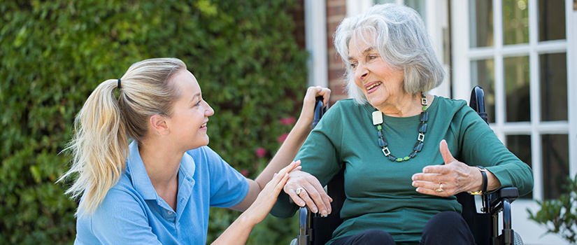 Image of a caretaker kneeling in front of a patient in a wheelchair listening to her speak.