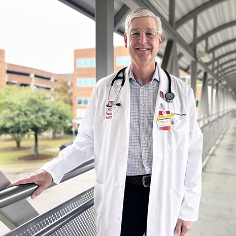 Headshot of Rob McMahon in a white lab coat standing on a walking bridge between two buildings.