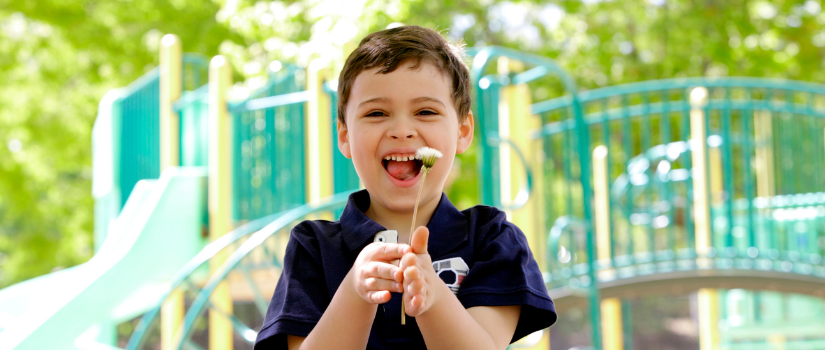 Young child smiling and holding a dandelion while playing on a playground.